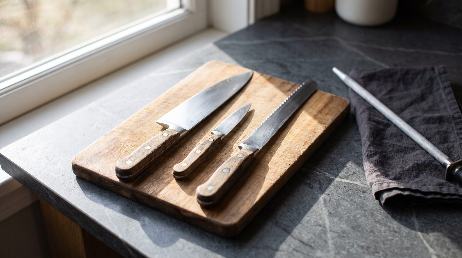 Three kitchen knives laid out on a wooden board with a honing steel beside them